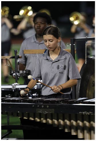 A person wearing a gray polo shirt with a band logo is playing a percussion instrument with mallets while other band members play brass instruments in the background. Descripci n generada con IA