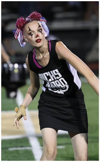 People in a marching band setting include one person playing a percussion instrument in the foreground, another playing a brass instrument in the background, and a person with colorful hair accessories and face paint performing on a field. Descripci n generada con IA
