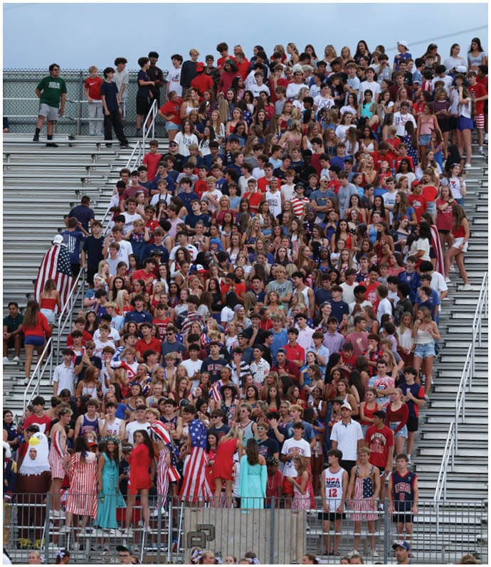 A large crowd of people wearing red, white, and blue clothing is gathered closely together on bleachers at an outdoor event, with some people standing on the steps and a few others walking nearby. Descripci n generada con IA