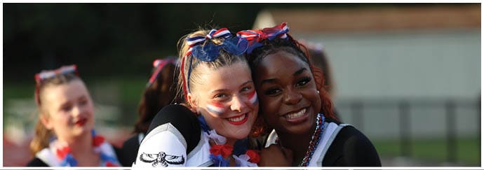 Two people wearing cheerleading uniforms and festive accessories smile and embrace outdoors with others in similar attire in the background. Descripci n generada con IA