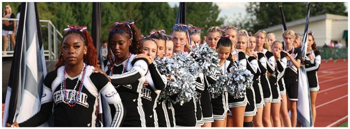 A line of people dressed in black and white cheerleading uniforms stand on a track field holding pom-poms and flags. Descripci n generada con IA