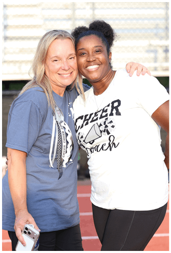 Two adults smiling and standing close together on a sports track with one wearing a shirt that says “CHEER coach." Descripci n generada con IA