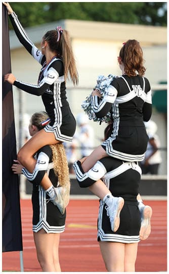 Two people in black and white cheerleading uniforms are holding a large banner, with one person sitting on the other's shoulders while reaching up to adjust the banner. Descripci n generada con IA