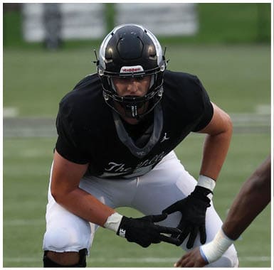 A person wearing a black helmet, black shirt, white pants, and a knee brace is crouched in a ready position on a football field with two other players partially visible nearby. Descripci n generada con IA