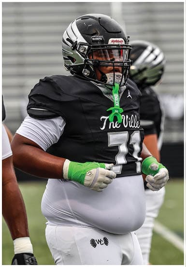 A person wearing a black football helmet and jersey with a green mouthguard hanging from the face mask is focused on the game. Descripci n generada con IA