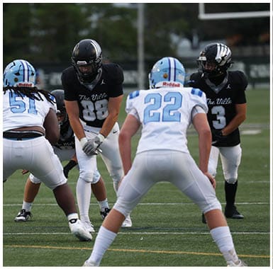 Four people in football uniforms are positioned on a field with two wearing black jerseys and two wearing white jerseys, appearing to prepare for a play. Descripci n generada con IA