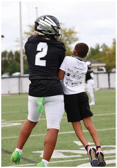 An adult in a black football uniform with the number two is holding a child wearing a white t-shirt and a beaded necklace on a sports field. Descripci n generada con IA
