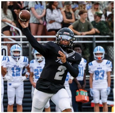 A person in a black football uniform is throwing a ball while several people in white and blue football uniforms stand in the background near a fence. Descripci n generada con IA