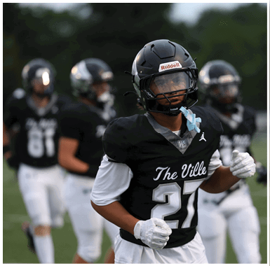Three people wearing black football uniforms and helmets stand on a field, with one person in the foreground looking to the side. Descripci n generada con IA