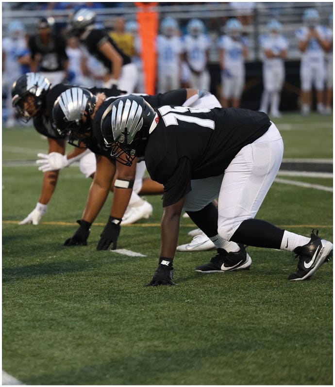 Two people in football uniforms and helmets are crouched in a ready position on a grassy field, preparing for a play. Descripci n generada con IA