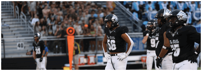 Several people in black football uniforms and helmets stand on a field with a crowd in the background. Descripci n generada con IA