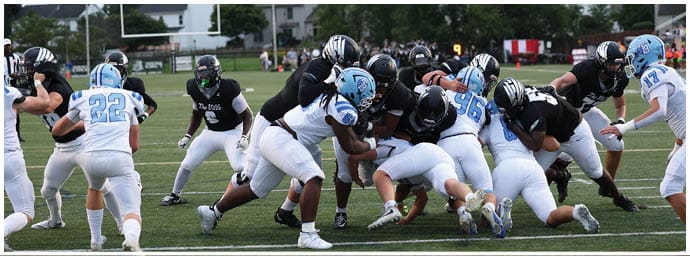 People in football uniforms are engaged in a play on a grassy field with houses and trees in the background. Descripci n generada con IA
