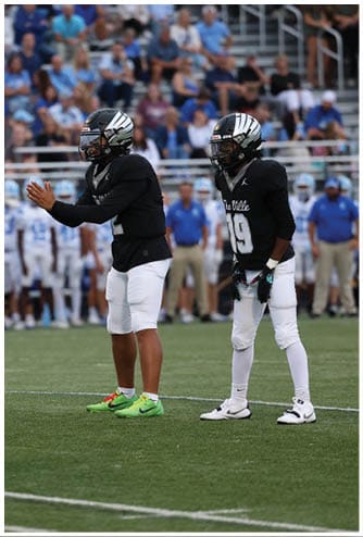 Two people wearing black football jerseys, white pants, and helmets stand on a football field with a blurred crowd in the background. Descripci n generada con IA