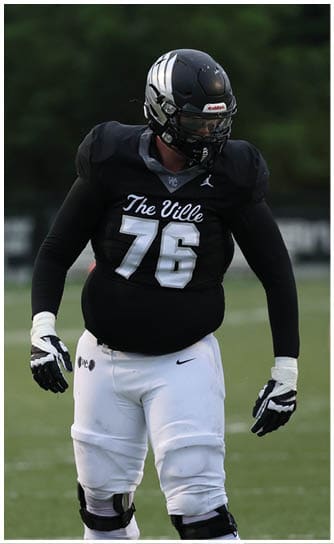 A person wearing a black football jersey with the number 76 and white pants stands on a green football field. Descripci n generada con IA