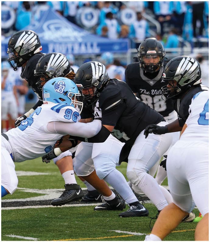 Several football players in black and white uniforms are engaged in a close, intense play on the field with one player in a white and blue uniform pushing against them. Descripci n generada con IA