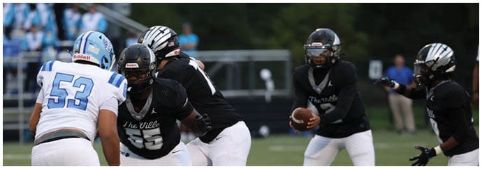 People in football uniforms are engaged in a play on a field with a crowd visible in the background. Descripci n generada con IA