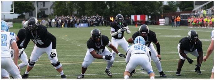 People in football uniforms are positioned on a field preparing for a play during a game. Descripci n generada con IA