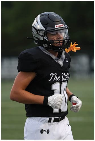 A person wearing a black football uniform and helmet is standing on a field with a small orange hand-shaped mouthpiece attached to the helmet. Descripci n generada con IA