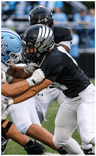 Two people wearing black football uniforms and helmets are engaged in a physical play on a football field. Descripci n generada con IA
