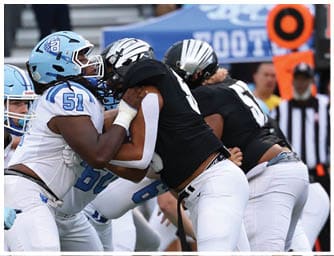 People wearing light blue and white football uniforms are engaged in a play on the field with one person holding another in a black uniform. Descripci n generada con IA