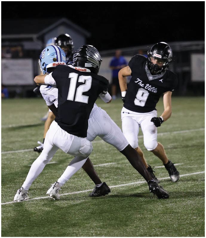 Two people in football uniforms are engaged in a tackle on a grassy field during a nighttime game. Descripci n generada con IA