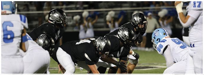 Adults in football uniforms are lined up on a field facing each other, preparing for a play during a game. Descripci n generada con IA