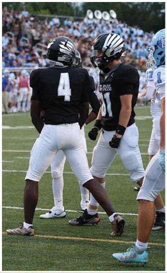 A person wearing a black football jersey with the number 4 and white pants stands on a football field with other players and a crowd in the background. Descripci n generada con IA