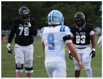 A person wearing a black football jersey with the number 63 and white pants stands on a football field between two other people in white and blue football uniforms. Descripci n generada con IA
