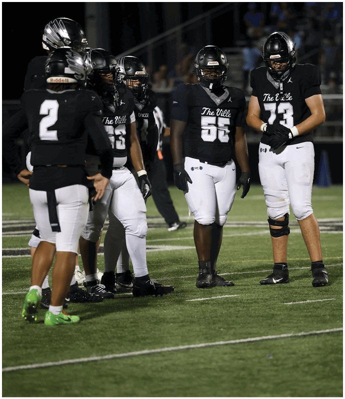 Three people wearing black helmets, black jerseys with “The Ville" text, and white pants stand on a football field during a game. Descripci n generada con IA