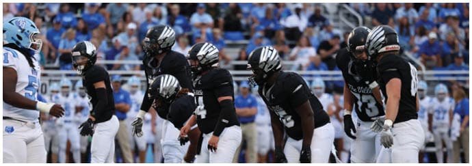 A group of people wearing black and white football uniforms are lined up on a field, preparing for a play during a game with spectators in the background. Descripci n generada con IA