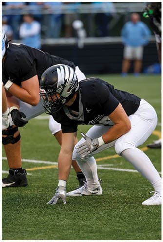 Two people wearing black and white football helmets and uniforms are engaged in a play on a football field. Descripci n generada con IA