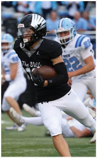 A person wearing a black football jersey with “The Ville" written on it is running while holding a football, with other players in white uniforms in the background. Descripci n generada con IA