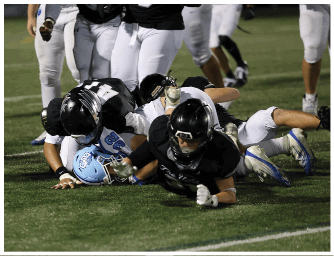 Several people in football uniforms are on a grassy field with one person in a light blue helmet being tackled by others in black helmets and black jerseys. Descripci n generada con IA