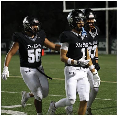 Three people wearing black football helmets and black jerseys with white pants are standing on a football field at night. Descripci n generada con IA