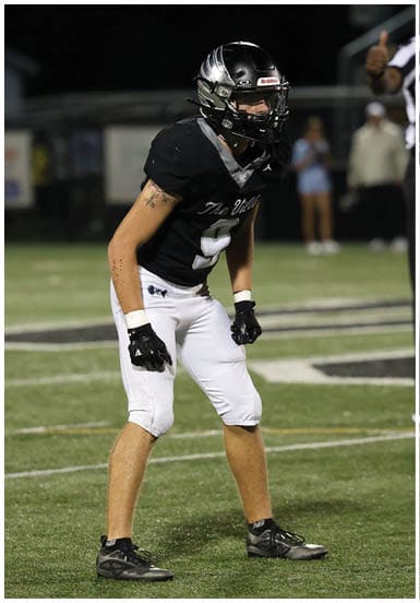 A person wearing a black football helmet, black jersey, white pants, and black gloves is standing in a ready position on a football field at night with a referee in the background. Descripci n generada con IA