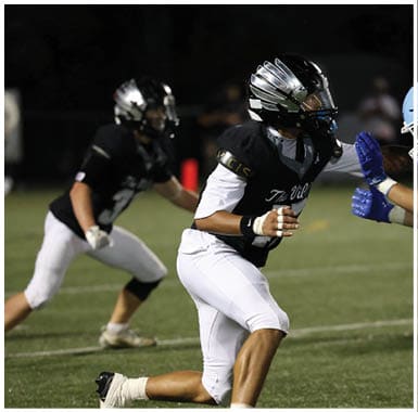 Two people wearing football helmets and uniforms are engaged in a play on a football field at night. Descripci n generada con IA