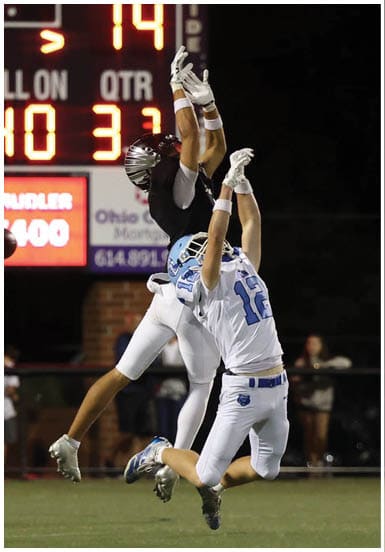 Two people in football uniforms jump with arms extended attempting to catch a football near a scoreboard at night. Descripci n generada con IA