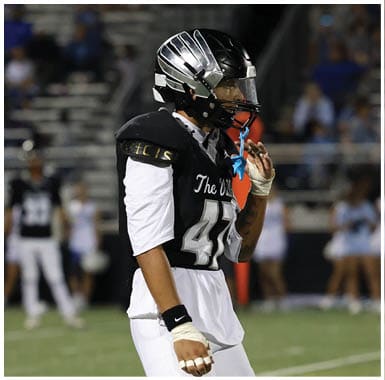 A person wearing a black and white football uniform and helmet stands on a field with blurred spectators and cheerleaders in the background. Descripci n generada con IA