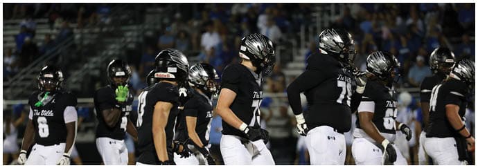 A group of people wearing black football uniforms and helmets are standing on a field with a crowd in the background. Descripci n generada con IA