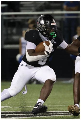 Two people in black football uniforms are running with the ball while two people in white and blue uniforms prepare to block or tackle on a football field. Descripci n generada con IA