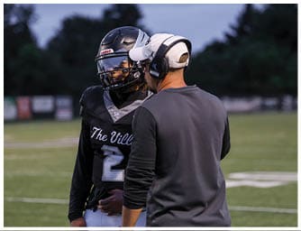 A person wearing a football helmet and uniform is standing on a field talking to another person wearing a headset and dark clothing. Descripci n generada con IA