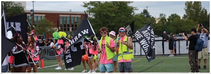 People wearing bright pink shirts and yellow vests are holding black flags with white text while standing on a sports field with other people and photographers in the background. Descripci n generada con IA
