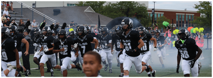 A group of people wearing black and white football uniforms and helmets are running onto a field through smoke with a crowd and cheerleaders holding yellow and green pom-poms in the background. Descripci n generada con IA