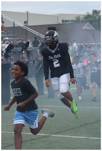 A person wearing a black t-shirt and blue shorts is running and smiling on a sports field while another person in a black football uniform and helmet stands nearby with a marching band playing in the background. Descripci n generada con IA