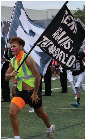 A group of people wearing black helmets and sports uniforms are running onto a football field with a person in a neon vest holding a flag nearby. Descripci n generada con IA