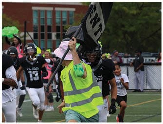 A group of people wearing black helmets and black and white sports uniforms are running on a football field with some holding large black flags while a person in a neon yellow vest runs alongside them. Descripci n generada con IA