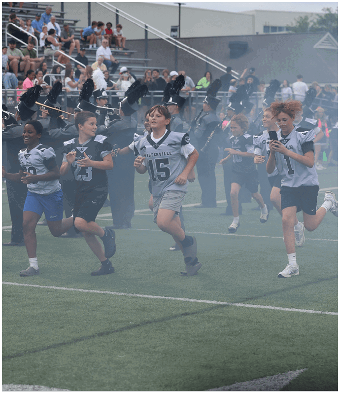 Children in sports uniforms are running on a field with a marching band playing in the background and spectators watching from the stands. Descripci n generada con IA