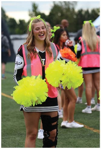 A person wearing a neon pink vest and a knee brace is smiling and holding bright yellow pom-poms on a sports field with other cheerleaders in the background. Descripci n generada con IA