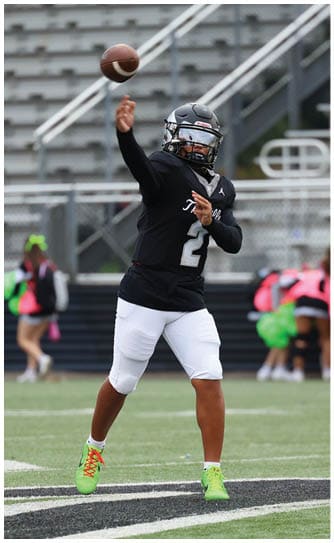 A person wearing a black Hawks shirt and gray pants stands on a sports field with arms crossed. Descripci n generada con IA