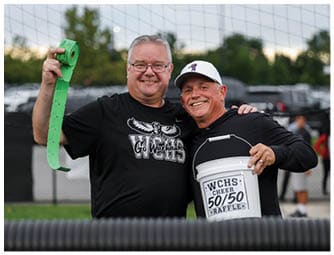Two adults smiling and posing closely together outdoors, one wearing glasses and a black shirt with a bird graphic, and the other wearing a white cap and a black shirt. Descripci n generada con IA
